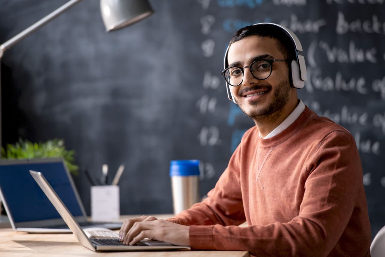 Portrait of a male student wearing glasses and headphones, smiling while using a laptop with a blurred classroom chalkboard background