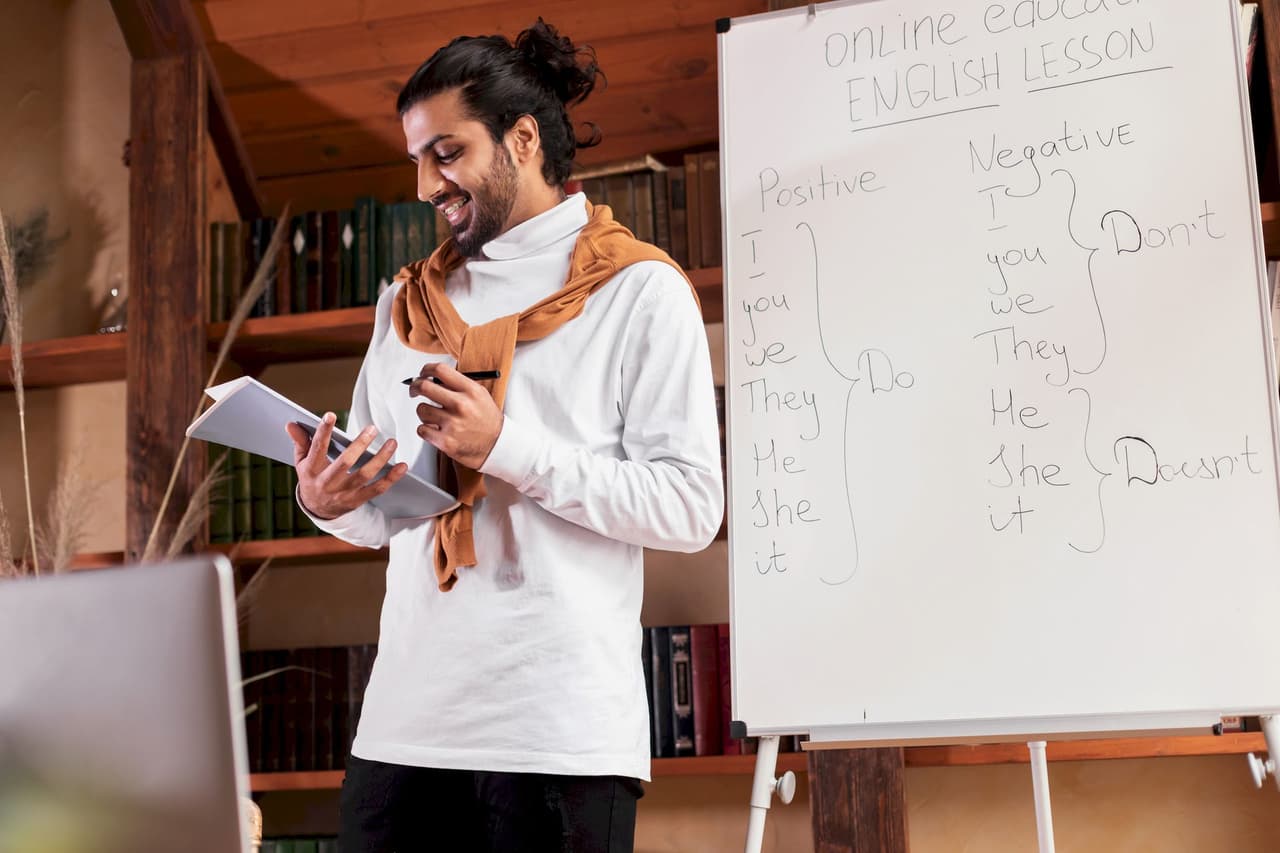 Male teacher conducting an online English lesson, standing next to a whiteboard with positive and negative sentence examples, in a cozy room with wooden walls and bookshelves.