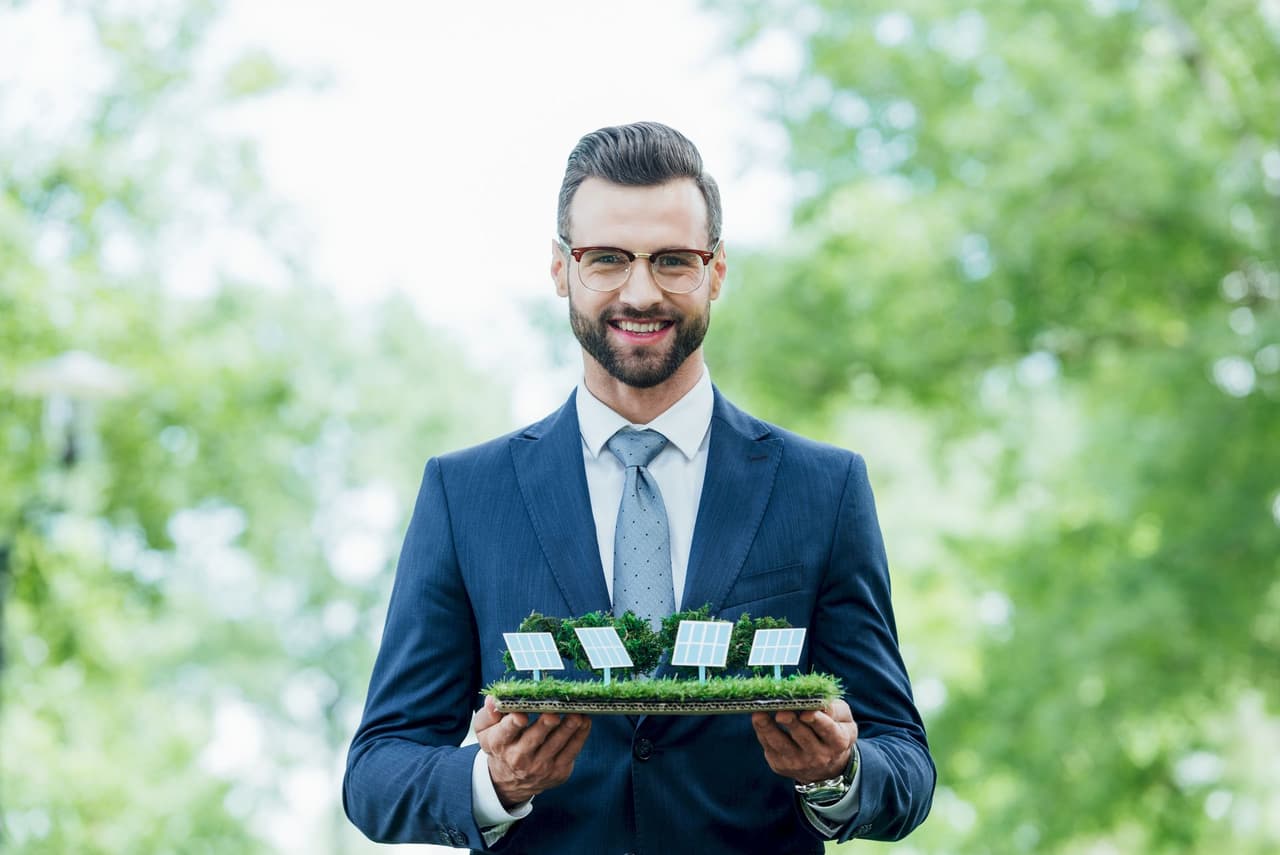 A smiling man in a blue suit and glasses holding a model of solar panels with a blurred green outdoor background