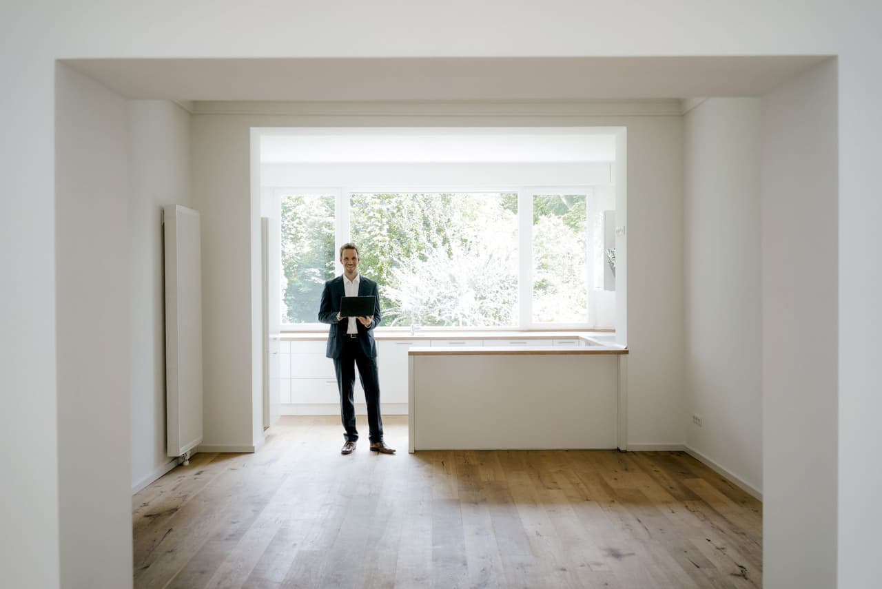 Man standing in an empty room near a large window with natural light and wooden floor
