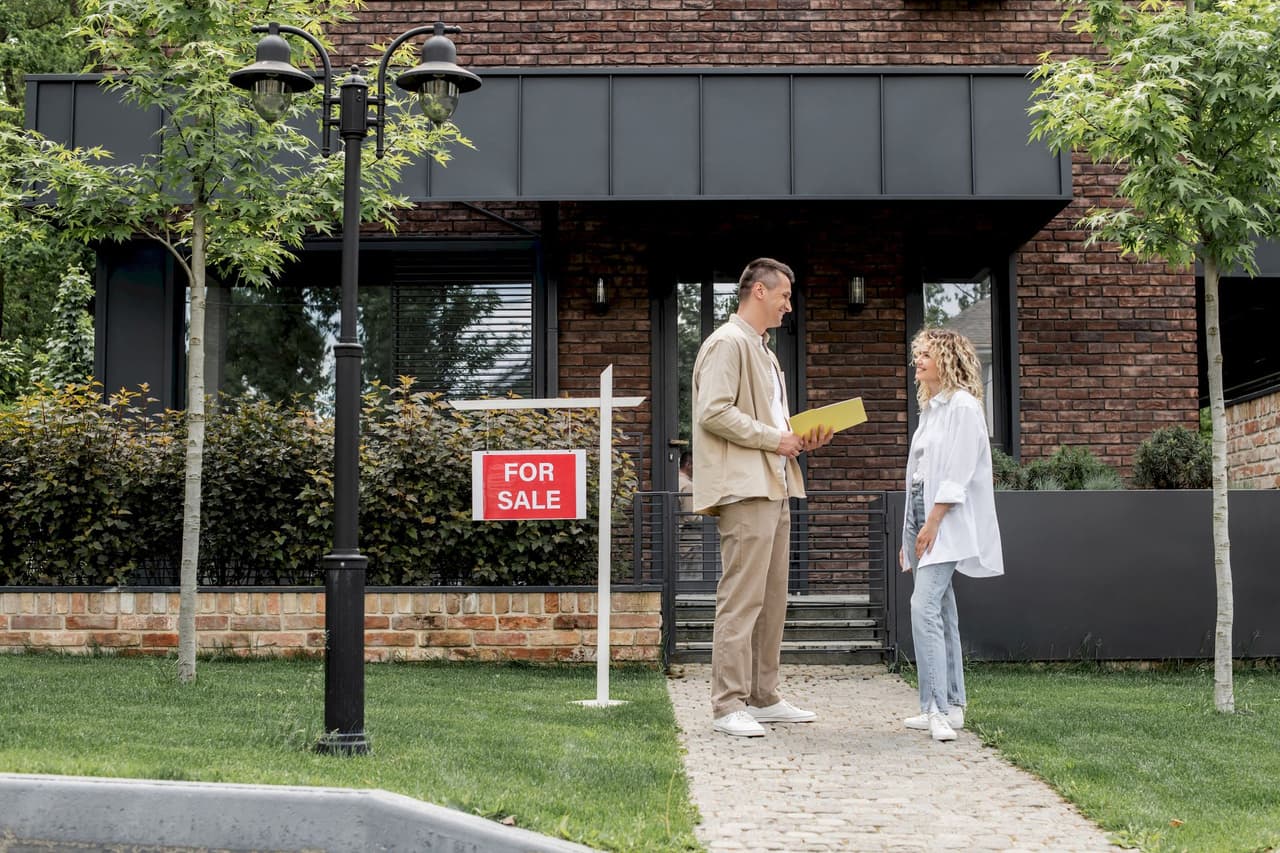 A man and a woman standing on a pathway in front of a house with a 'For Sale' sign, engaged in conversation, with greenery and a brick house in the background.