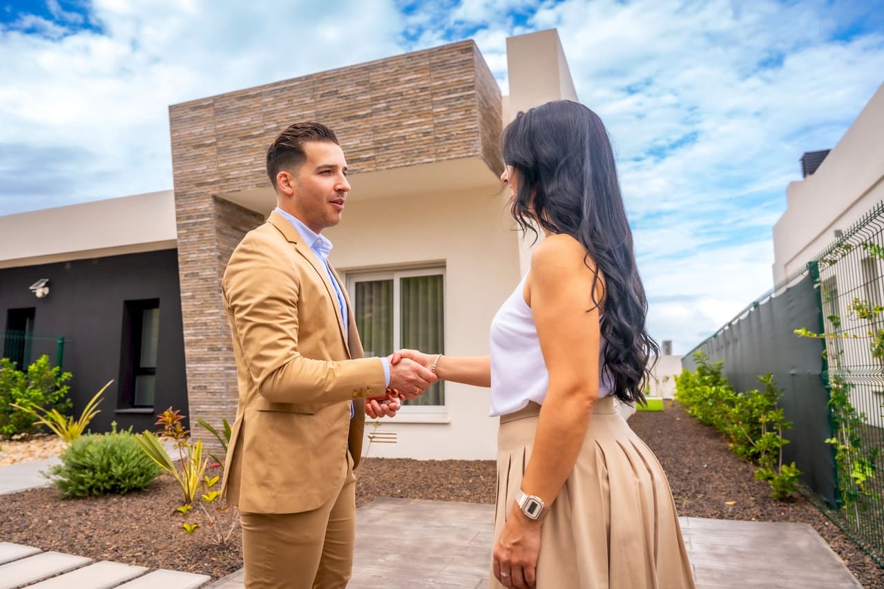 A man and a woman shaking hands outdoors in front of a modern house with a partly cloudy sky in the background.
