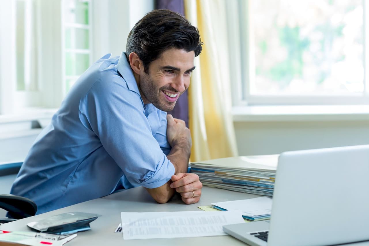Smiling man working at home office desk with laptop and documents