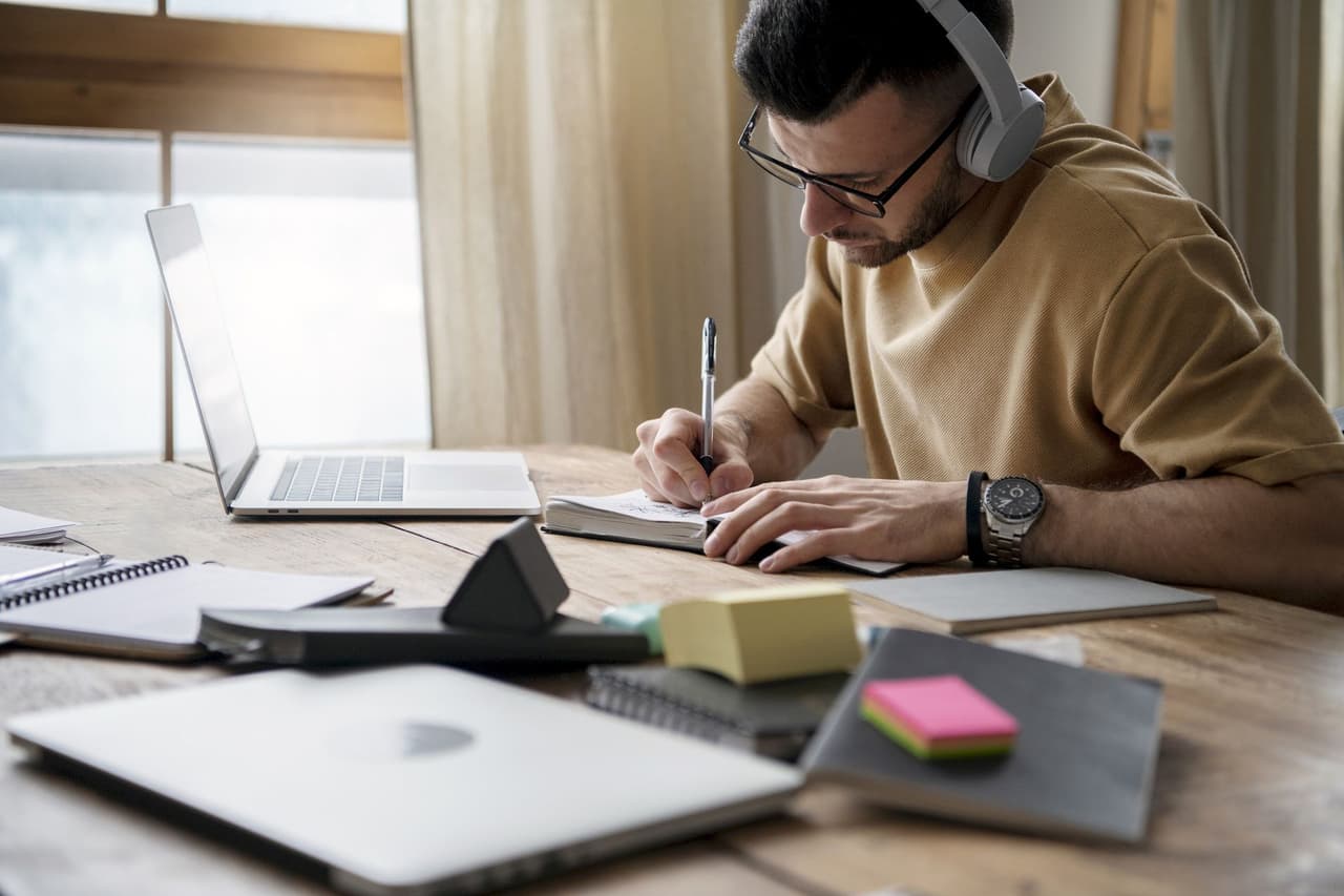 A man wearing headphones and glasses is writing notes in a notebook at a wooden desk with a laptop and office supplies around him in a well-lit room.