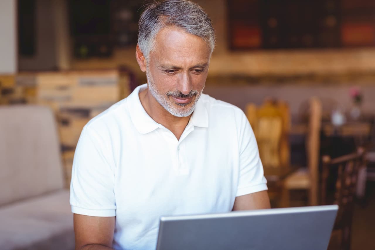 Middle-aged man with gray hair and beard wearing a white polo shirt, using a laptop in an indoor setting with a blurred background.