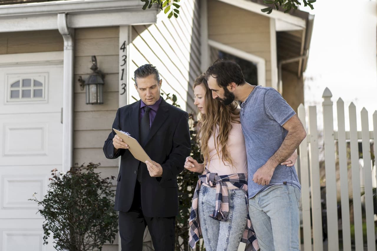 Real estate agent showing clipboard to a young couple outside a house with white picket fence