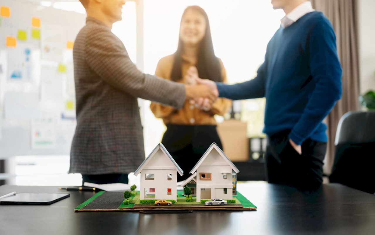 Two male real estate agents shaking hands with a female colleague clapping in the background, with model houses on the table in the foreground, office setting with bright natural light