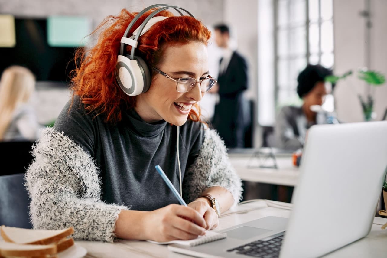 Redhead woman wearing headphones and glasses, smiling while writing in a notebook and looking at a laptop in a modern office with blurred background