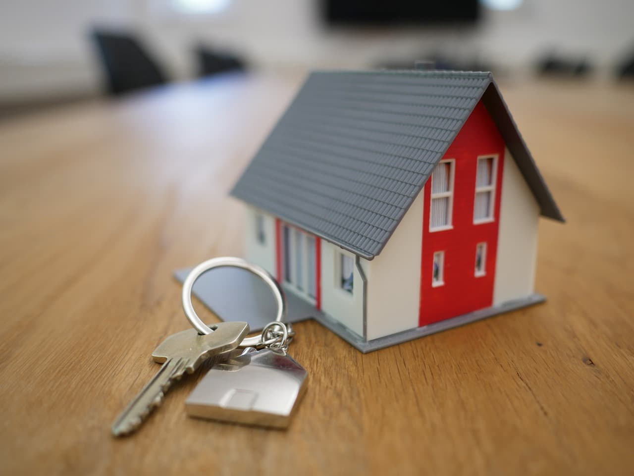 A small detailed model of a house with red and white walls and a grey roof placed on a wooden table, next to a keychain with two keys and a house-shaped pendant, with a blurred background