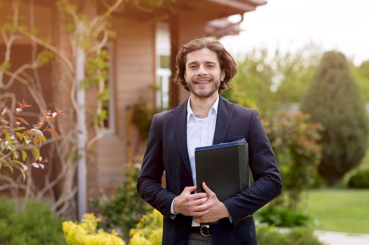 Portrait of a smiling male professional standing outdoors in a garden, holding a folder, with a blurred background of greenery and a wooden building.