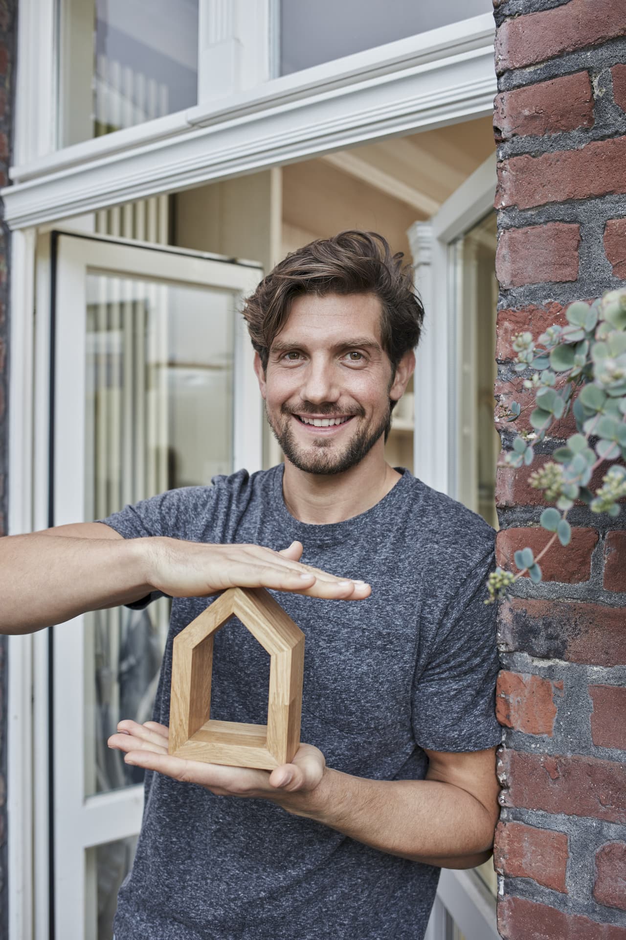 Smiling man with beard and short hair holding a small wooden house model in front of a brick wall and glass door, portrait style with clear background