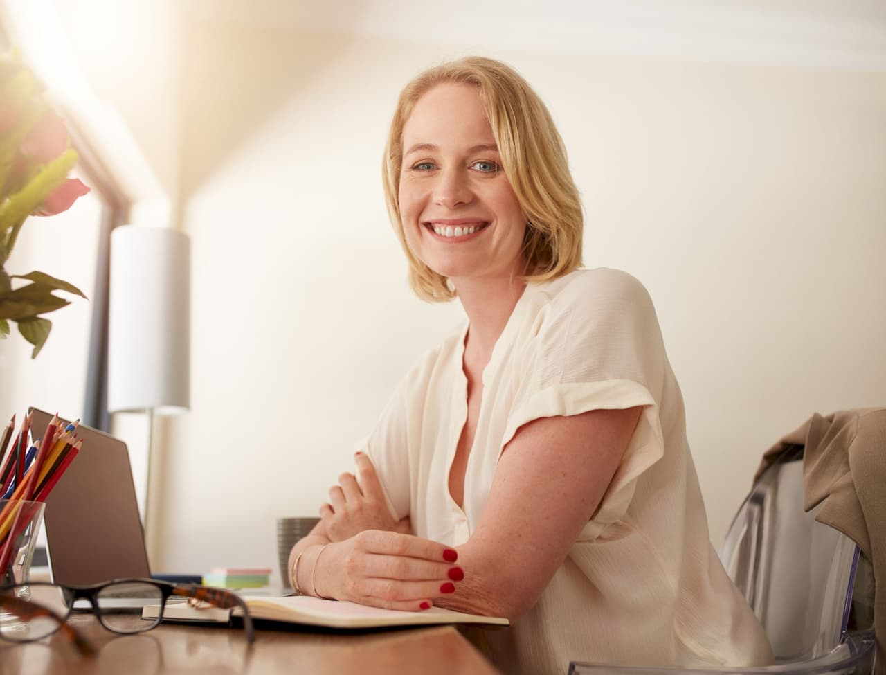 Portrait of a smiling woman sitting at an office desk with a laptop, notebook, and pencils, with a softly lit background.