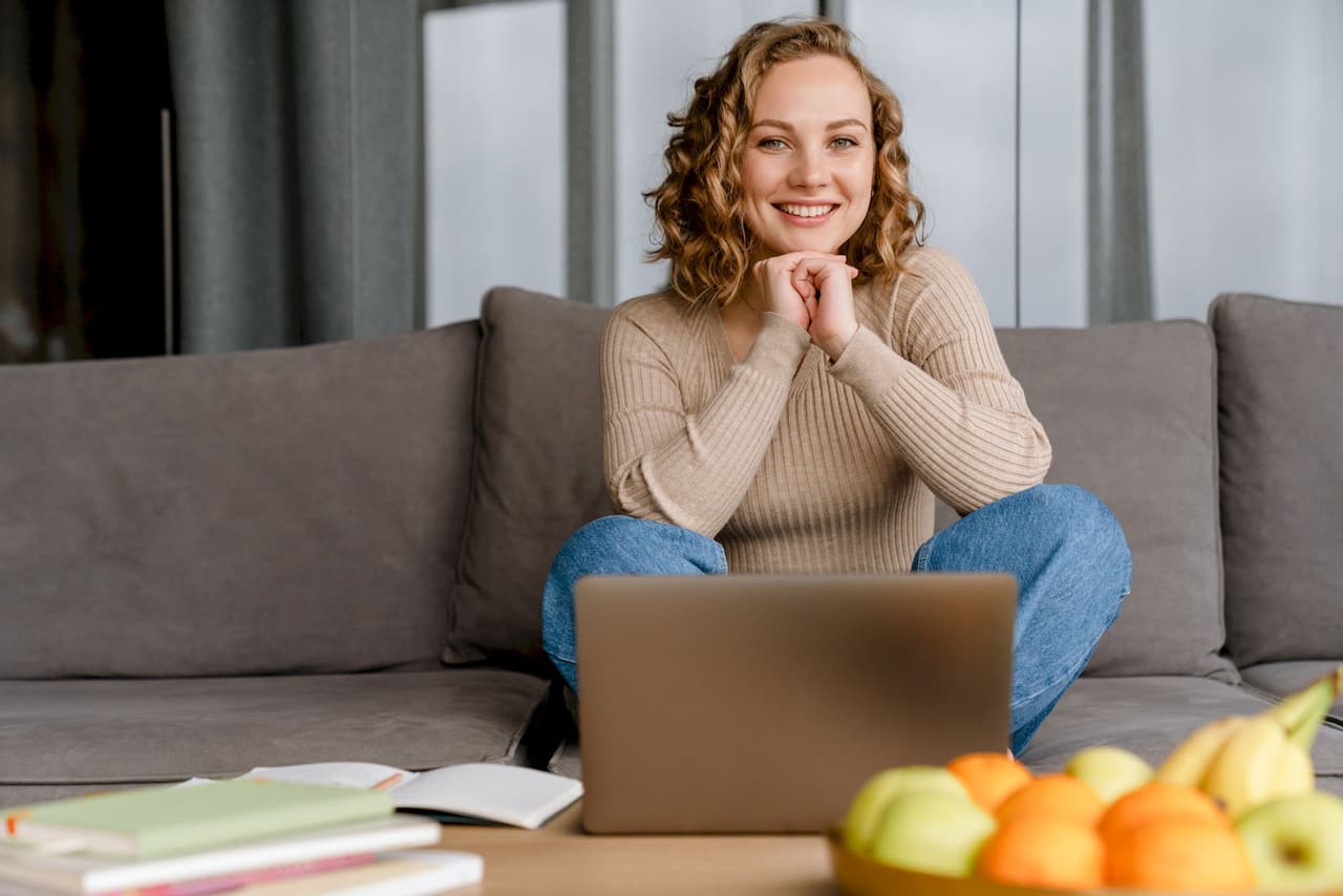 Smiling woman sitting cross-legged on a couch with a laptop in front of her, casual indoor setting with books and fruit on the table