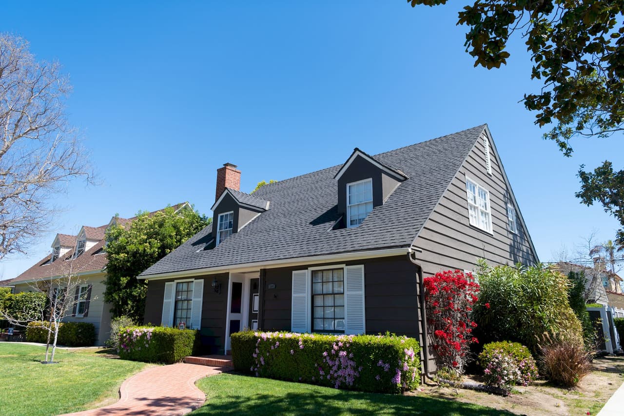 A suburban house with a dark gray roof and brown siding, surrounded by a garden with green bushes, red flowers, and a tree, under a clear blue sky during daytime.