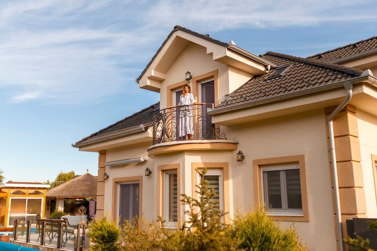 A woman standing on the balcony of a modern two-story house during daylight with clear blue sky in the background.