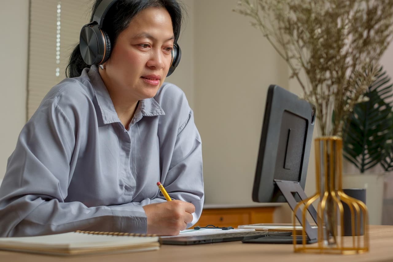 Woman wearing headphones sitting at a desk, taking notes while looking at a computer screen with a blurred background