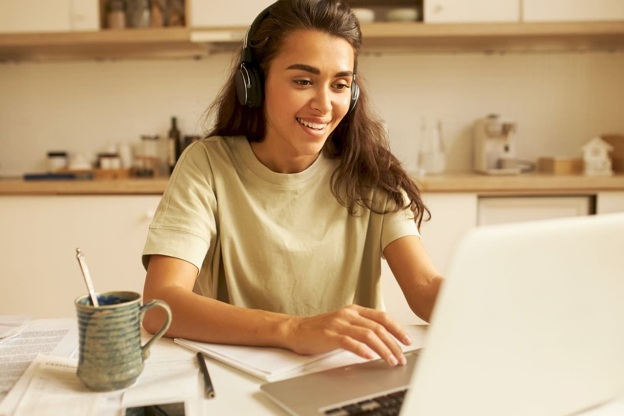 Young woman wearing headphones and smiling while using a laptop in a cozy kitchen setting with a blurred background