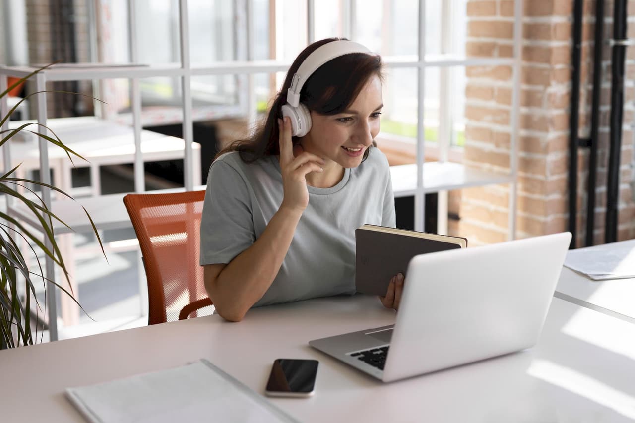 Young woman wearing headphones and holding a tablet while engaging in a video call on a laptop in a bright office setting
