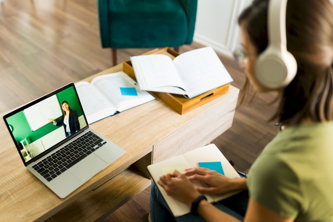 Woman with headphones attending an online class on a laptop, sitting at a wooden table with open books and notes