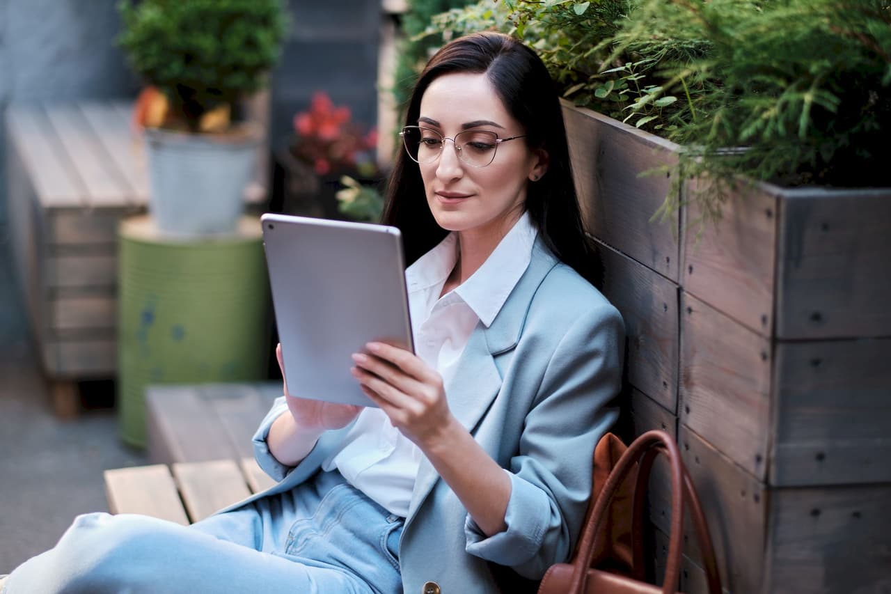 Portrait of a woman with glasses reading a tablet while sitting outdoors on wooden steps with plants in the background