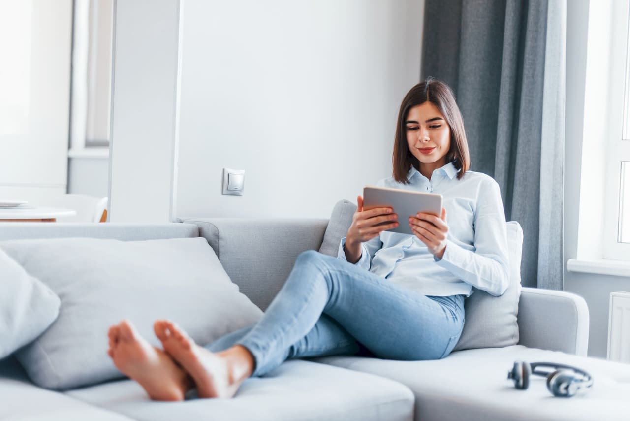 A young woman with dark hair sitting barefoot on a gray sofa, using a tablet in a bright living room with natural light coming through the window.