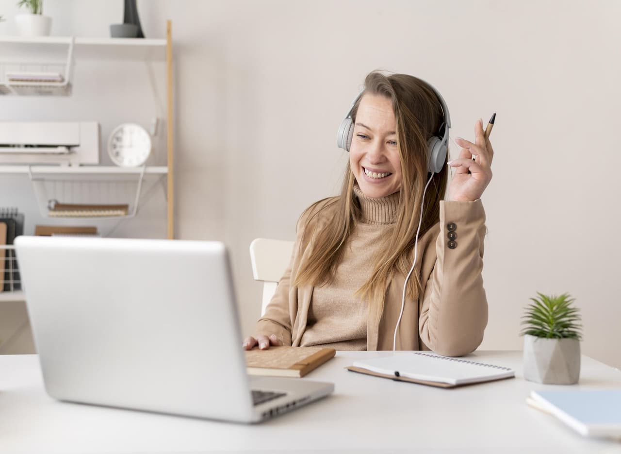 Smiling woman wearing headphones working remotely on a laptop at a desk with notebook and plant