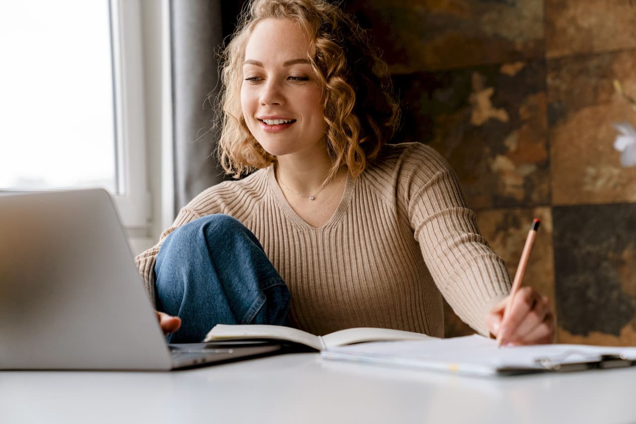 Young woman with curly hair studying and taking notes while looking at a laptop, sitting indoors with a blurred background