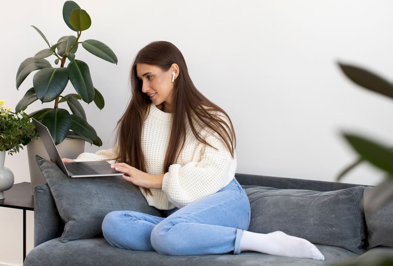 A young woman with long brown hair wearing a white sweater and blue jeans sitting cross-legged on a gray sofa, using a laptop. There are green plants in the background and the setting is indoors with a plain white wall.