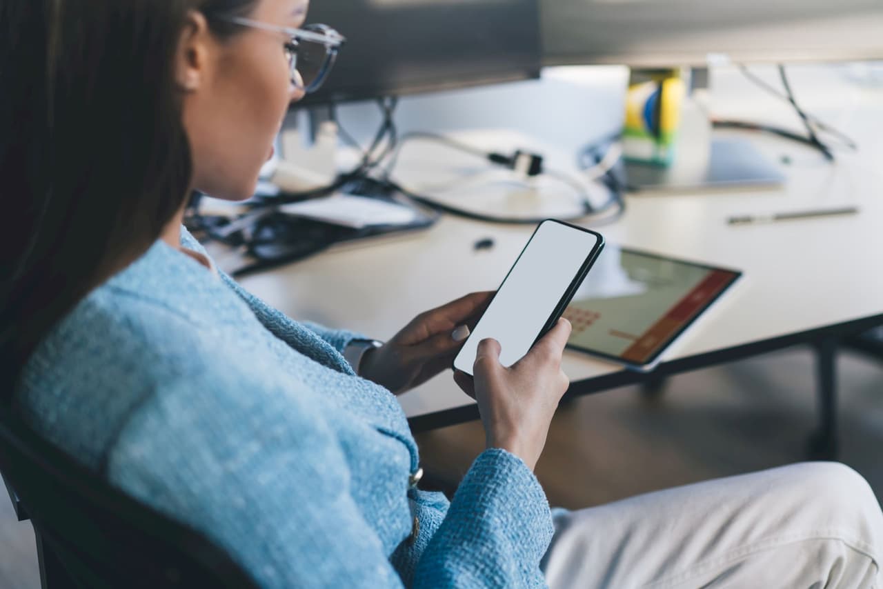 Portrait of a woman with glasses using a smartphone in an office setting with a blurred background and a tablet on the desk