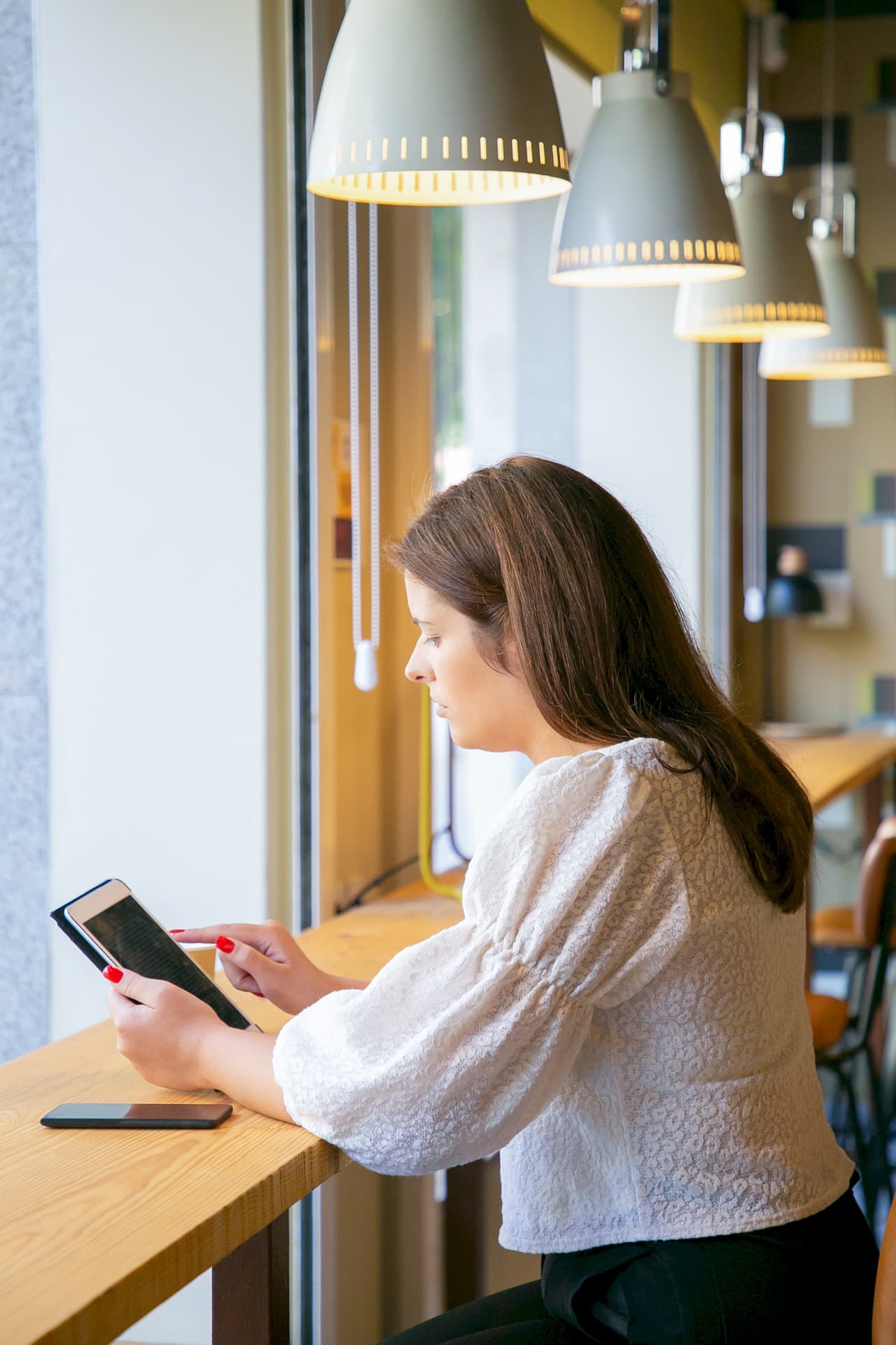 Side view portrait of a woman with long brown hair using a tablet while sitting at a wooden counter in a cafe with hanging lamps and a blurred background.