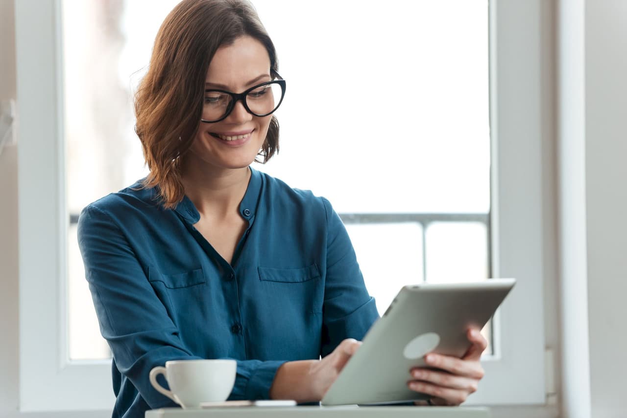 Smiling woman with glasses using a tablet in a casual indoor setting with a softly blurred window background