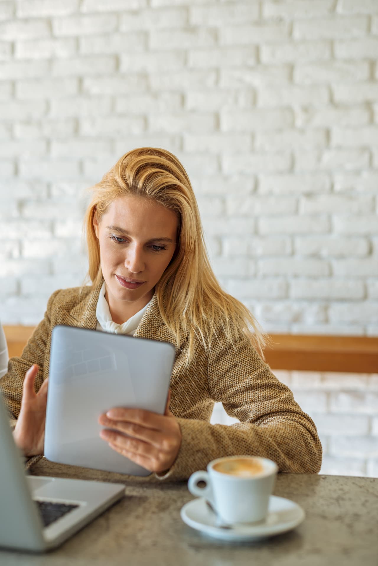 Young woman with blonde hair using a tablet in a coffee shop with a cup of coffee on the table and a laptop nearby, white brick wall background