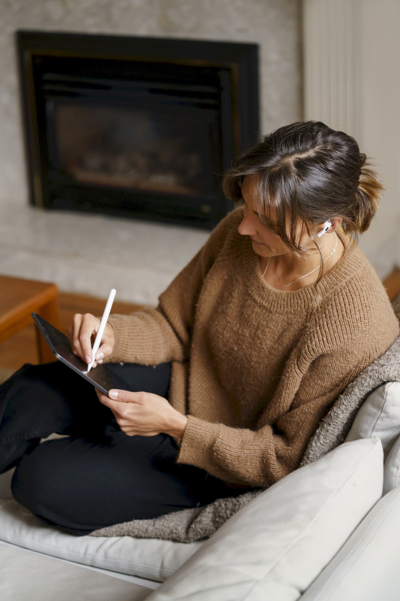 A woman with brown hair tied back, wearing a brown sweater and black pants, sitting cross-legged on a white couch in a cozy living room, using a tablet with a stylus. She is wearing wireless earbuds and there is a fireplace in the background.