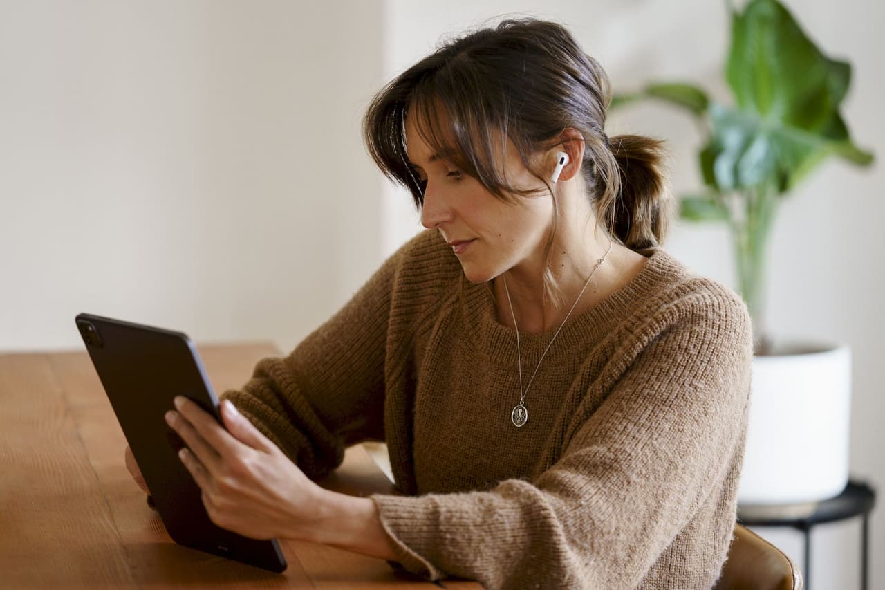 Portrait of a woman wearing earbuds and using a tablet while sitting indoors with a plant in the background