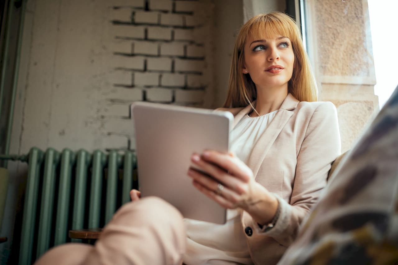 Portrait of a woman with blonde hair using a tablet indoors, sitting near a window with a blurred background of a radiator and brick wall.