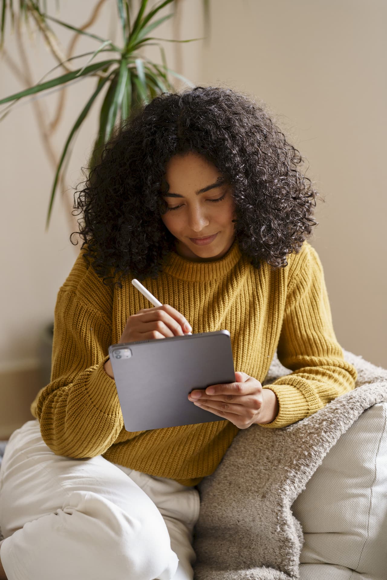 Young woman with curly hair wearing a mustard sweater, sitting on a couch and using a tablet with a stylus in an indoor setting with a plant in the background