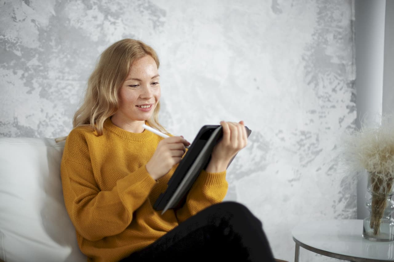 Young woman with blonde hair wearing a mustard sweater, sitting indoors on a white couch, using a tablet with a stylus, with a textured gray wall background in a portrait style