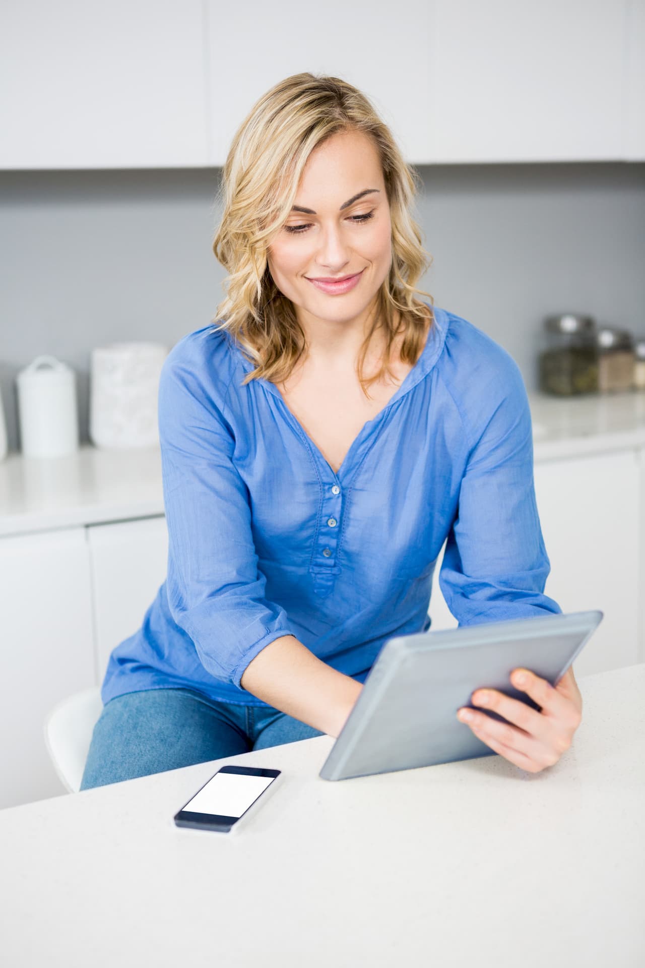 Portrait of a woman with blonde hair wearing a blue shirt, sitting at a kitchen counter and using a tablet, with a smartphone placed on the counter.
