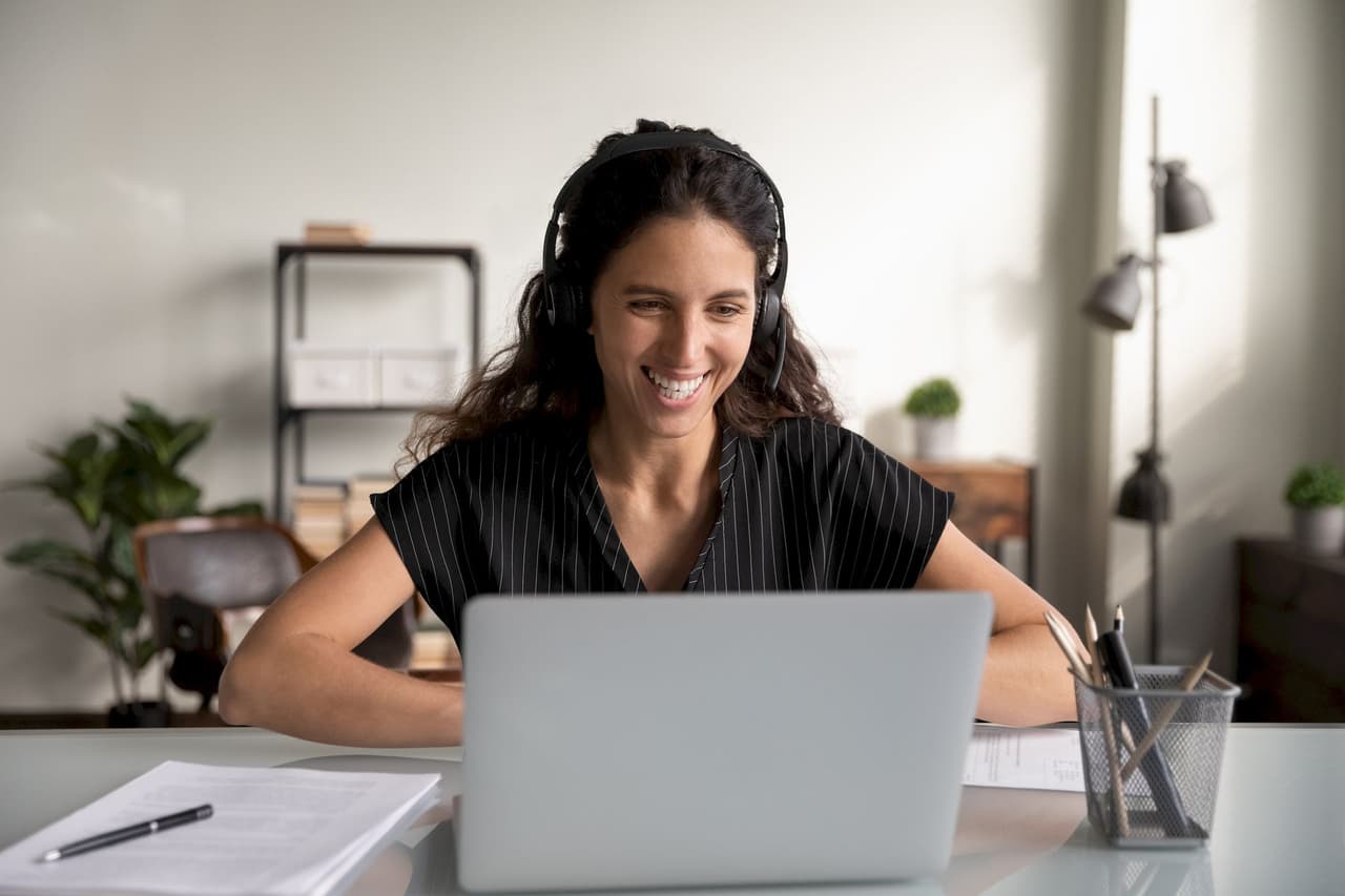 Smiling woman with headphones on video call using laptop in home office with blurred background
