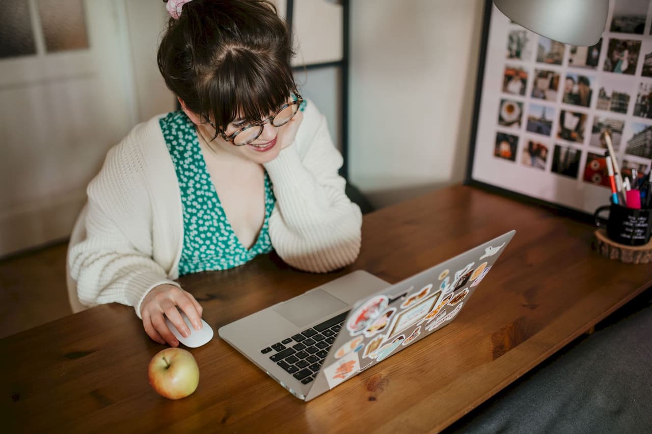 Young woman with glasses and a white cardigan working on a laptop at a wooden desk in a home office with a blurred background and an apple on the desk