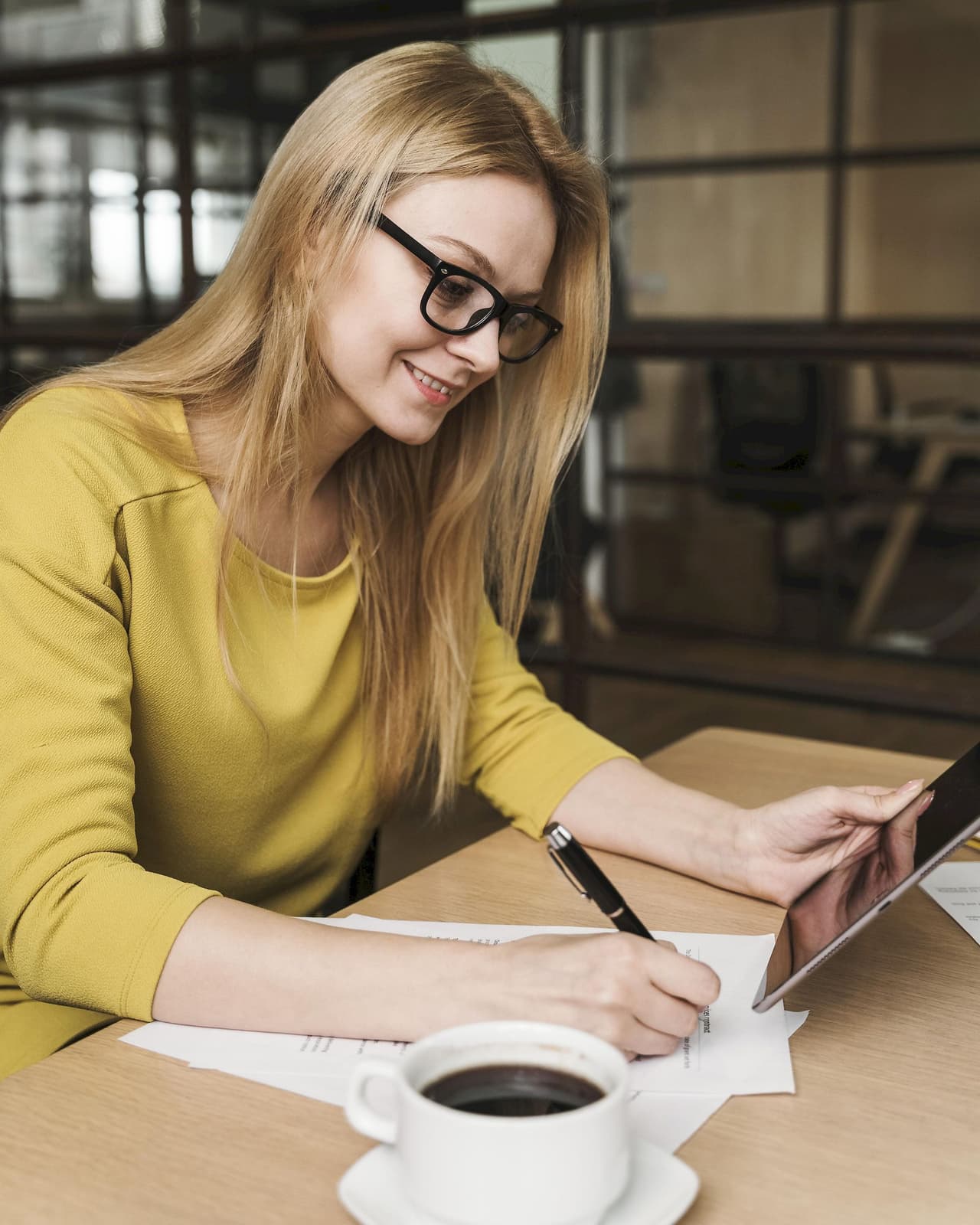 Young woman with glasses working on a tablet and writing on paper at a table in a cafe with a cup of coffee