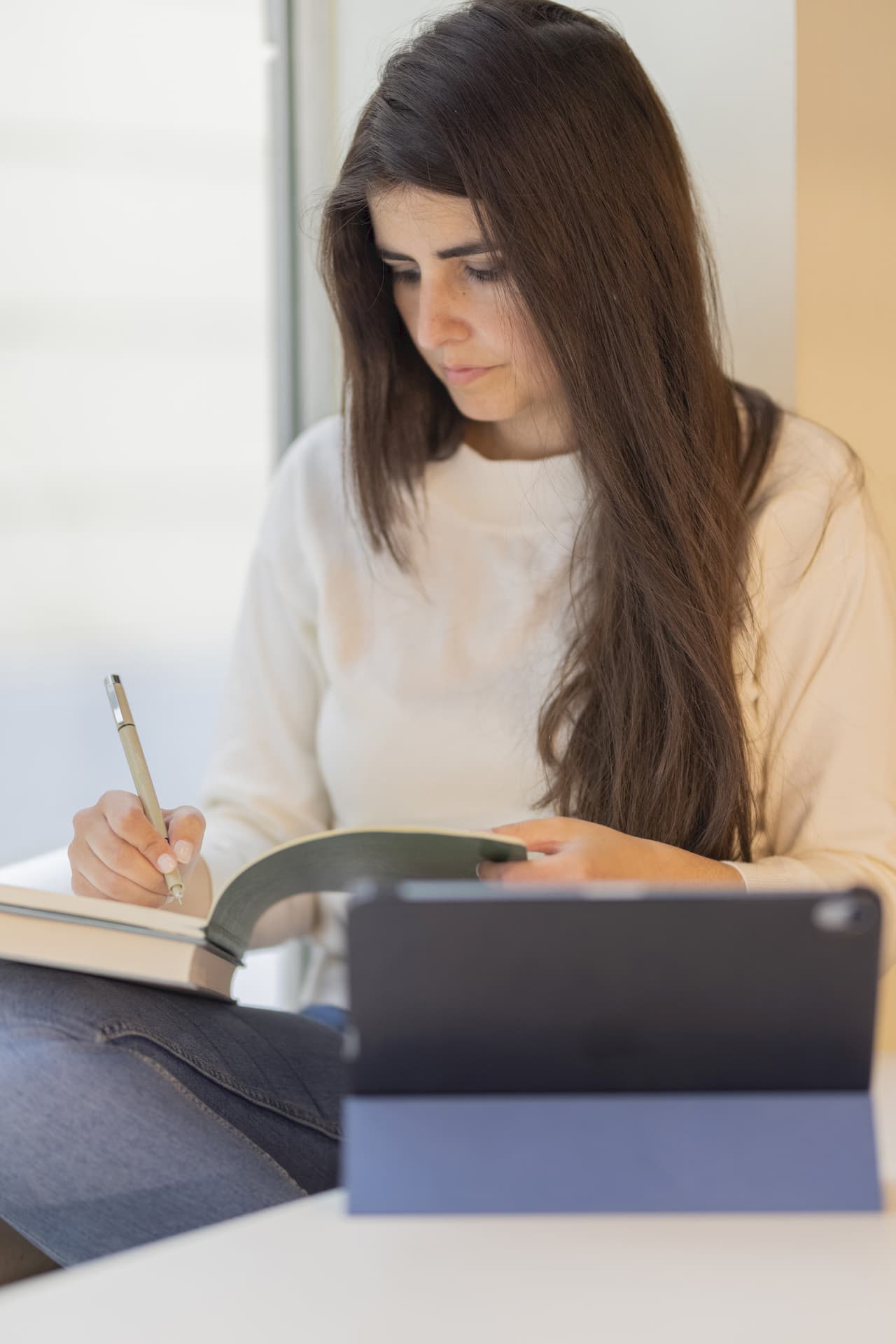 Portrait-style image of a woman with long dark hair writing notes in a notebook while sitting indoors with a tablet in front of her