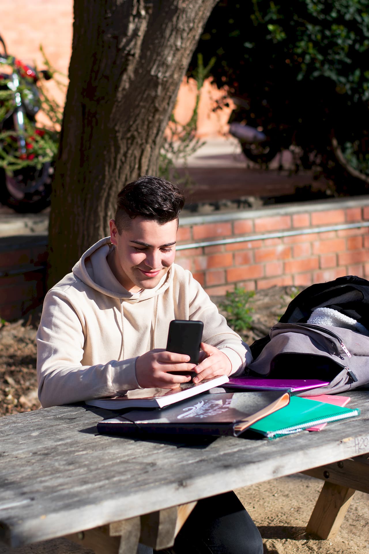 Young male student sitting at an outdoor wooden table, smiling and looking at his smartphone, with notebooks and a backpack on the table, and a blurred background of trees and a brick wall.