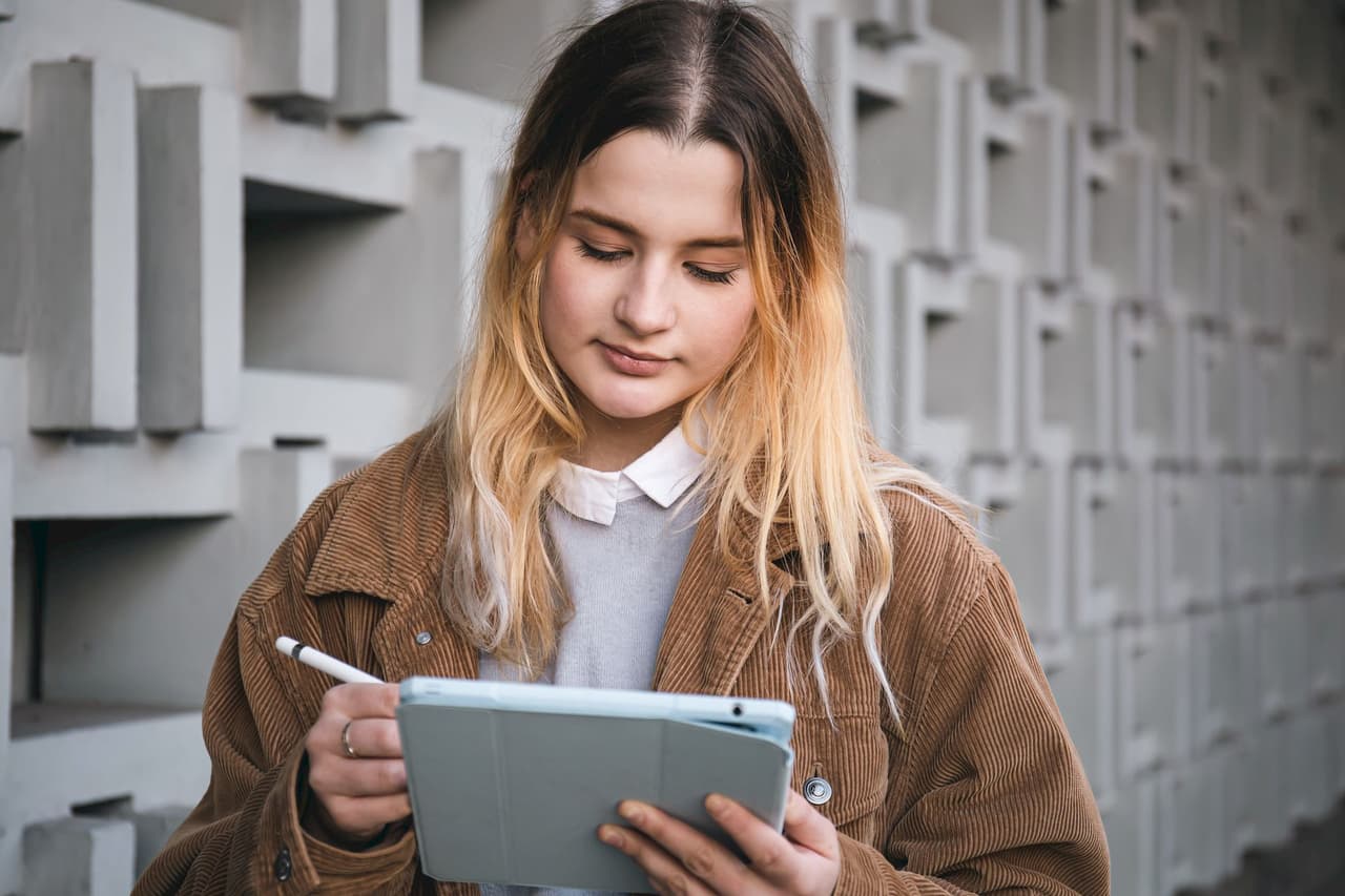 Young woman with long hair wearing a brown jacket using a tablet outdoors with a geometric concrete wall background